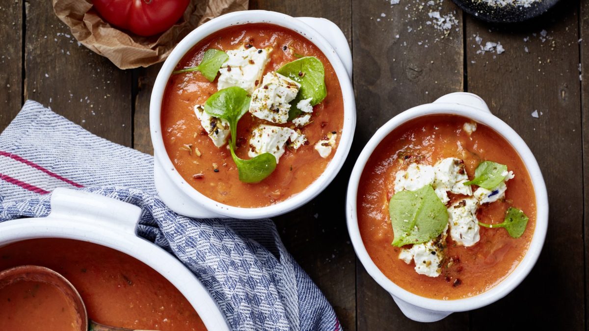 Zwei Schüsseln gefüllt mit Ofentomatensuppe mit mariniertem Deichkäse auf braunem Hintergrund. Links unten ein Topf mit Tomatensuppe und ein blaues Geschirrtuch.