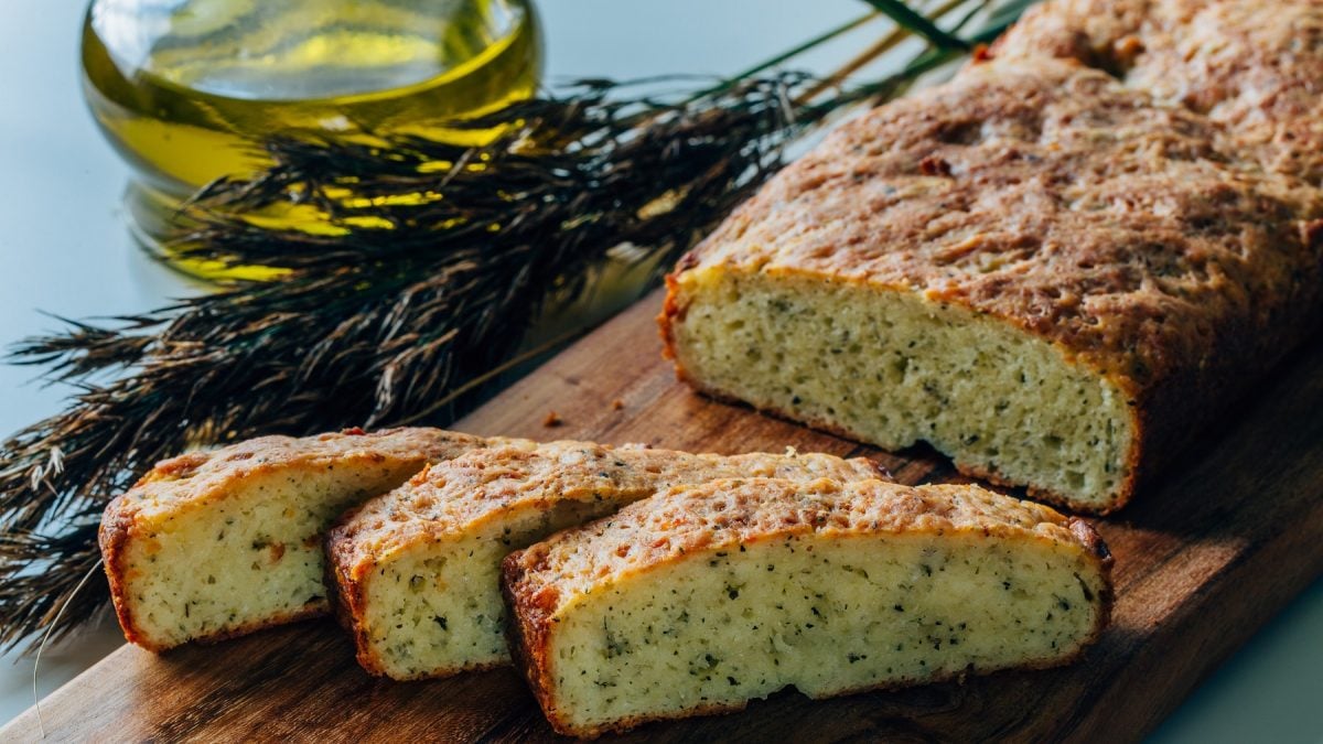Das Parmesanbrot angeschnitten auf einem rustikalen Holztisch mit einem grünen Strauch und Olivenöl im Hintergrund. Alles nah und von leicht schräg oben fotografiert.
