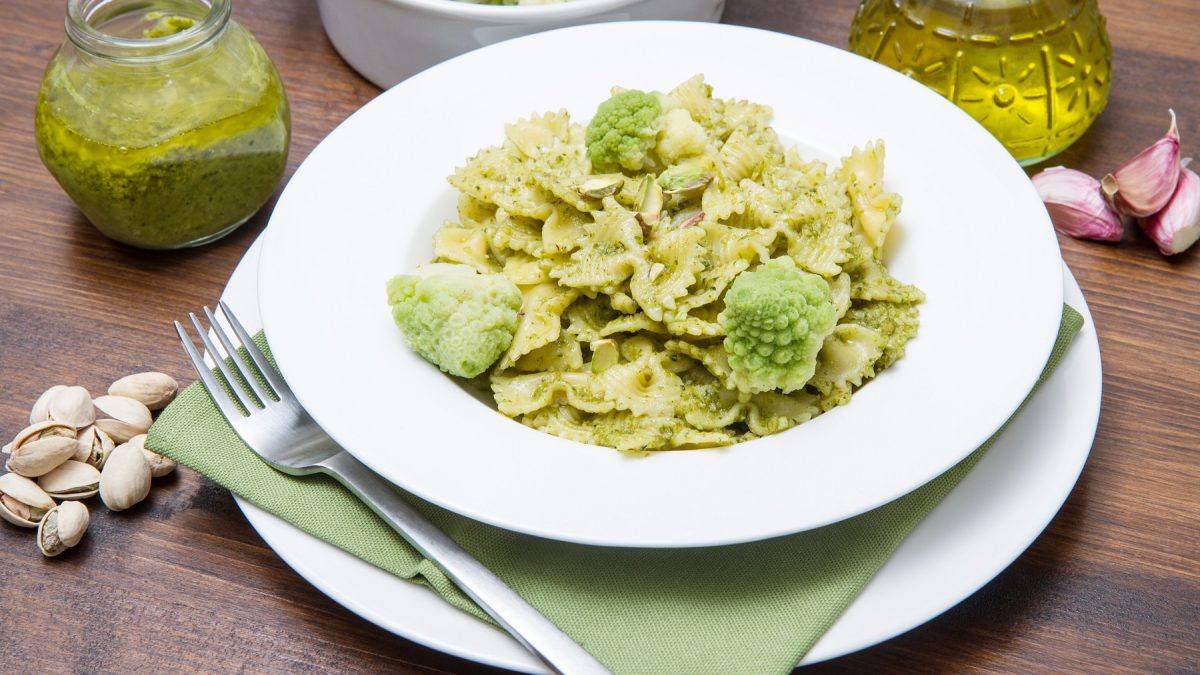 Ein weißer Teller und Unterteller mit einer hellgrünen Serviette und dem Pastagericht. Alles auf einem dunklen Holztisch vom schräg oben fotografiert. Drumherum Zutaten wie das Pesto, Brokkoli, Knoblauch und Olivenöl.