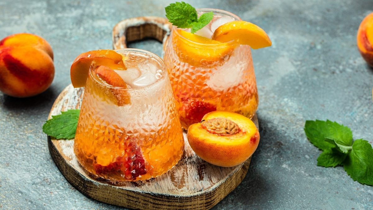 Pfirsich-Spritz-Bowle in zwei Gläsern auf einem Tablett mit angeschnittenem Pfirsich.