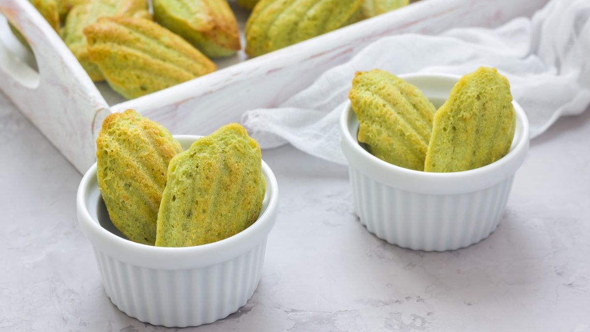 Einige Pistazien-Madeleines in kleinen weißen Schälchen und auf einem großen Brett im Hintergrund. Tisch, Schälchen und Untergrund sind in Grau- und Weißtönen gehalten und von leicht schräg oben fotografiert.