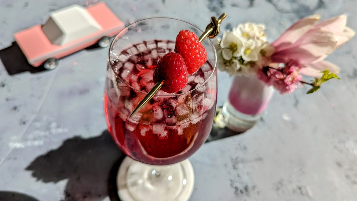 Ein Glas mit dem Cocktail auf grauem Steintisch. Unscharf einige Blüten und ein pinkes Holzauto im Hintergrund zu erkennen.