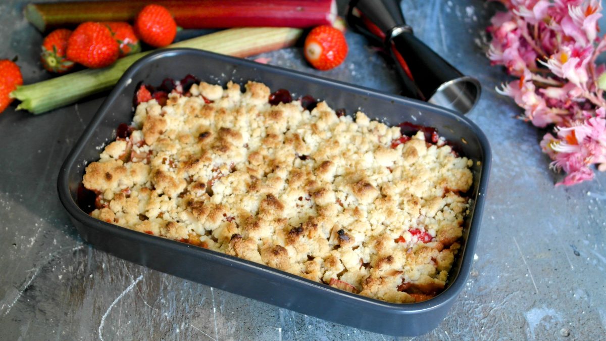 Eine graue Auflaufform mit dem Crumble auf einem grauen Steintisch. Dahinter pinke Blüten und einige Zutaten für das Dessert.