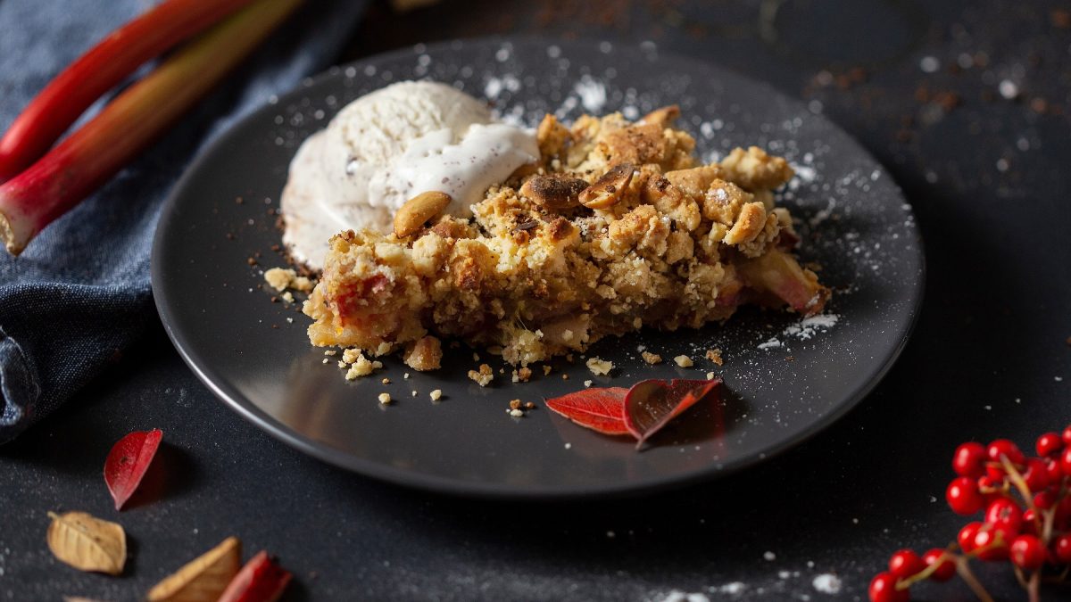 Ein dunkler Teller mit dem Crumble auf einem dunklen Tisch mit roter Deko und Rhabarberstangen.