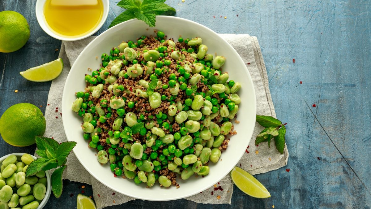 Saubohnensalat mit Quinoa und Erbsen in einer weißen Schüssel, daneben frische Minze, eine Schale mit Öl und Limetten.