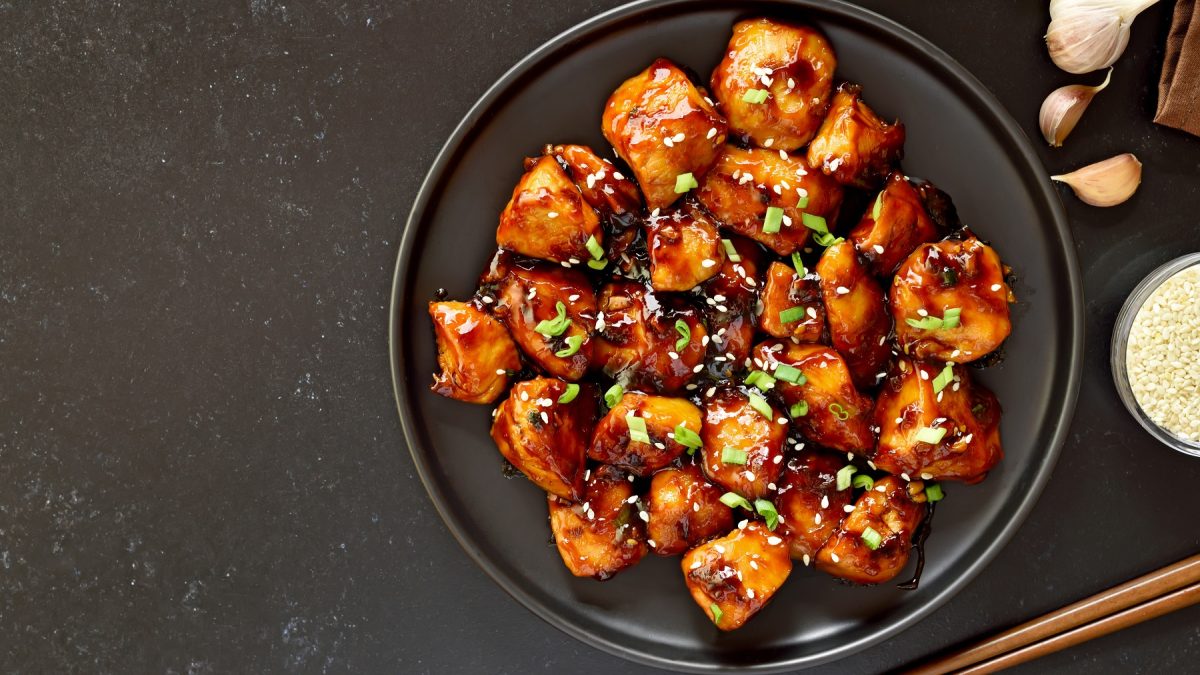 Ein schwarzer Teller mit dem Teriyaki-Hähnchen auf dunklem Untergrund. Rechts neben dem Teller diverse Zutaten für die Sauce.