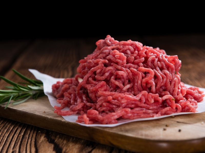 Rohes Hackfleisch auf Pergamentpapier und einem Holzbrett auf einem Holztisch von der Seite mit schwarzem Hintergrund fotografiert. Daneben noch ein Zweig Rosmarin.