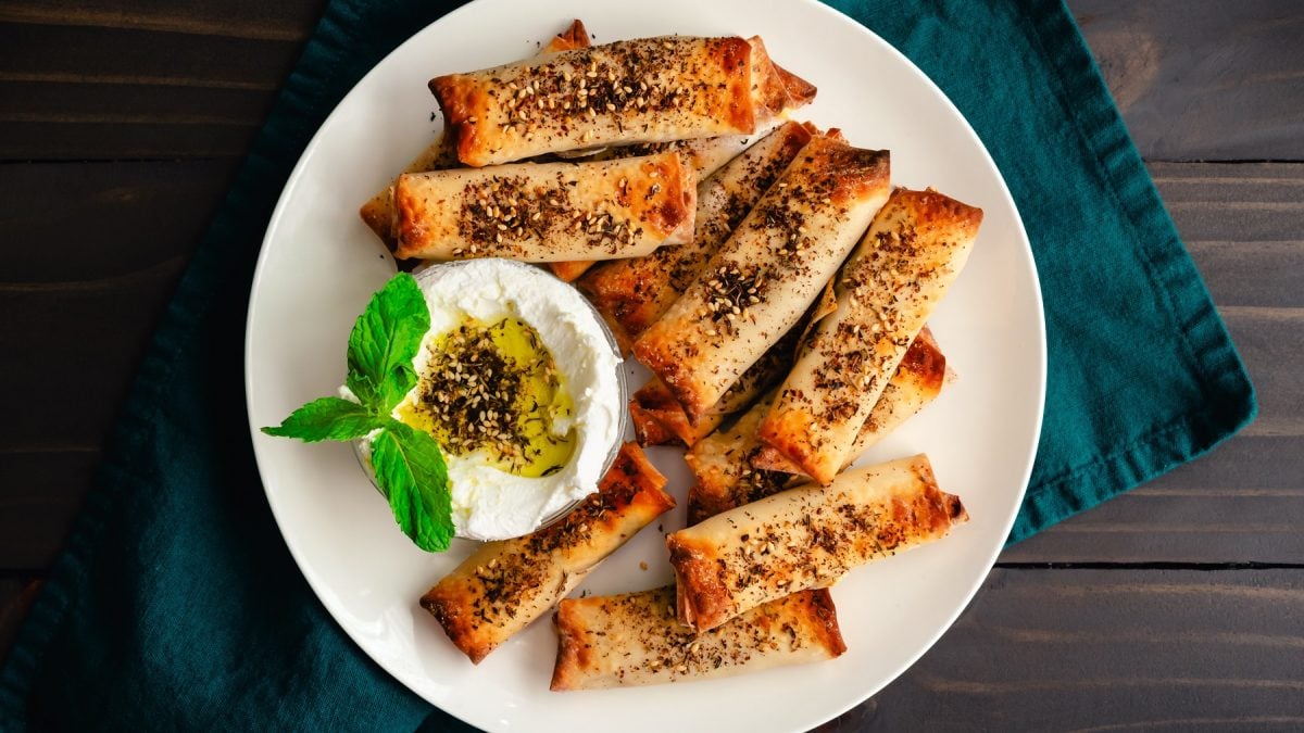 Za'atar-Halloumi-Röllchen mit einer Schale Labneh mit Olivenöl und Za'atar auf einem weißen Teller vor dunklem Hintergrund.