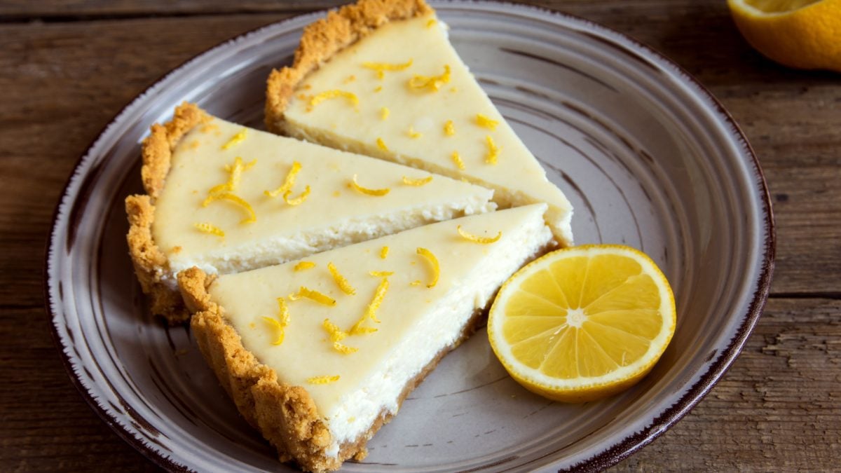 Ein grauer Teller mit drei Stücken vom Zitronen-Cheesecake auf einem rustikalen Holztisch. Alles von schräg oben fotografiert.