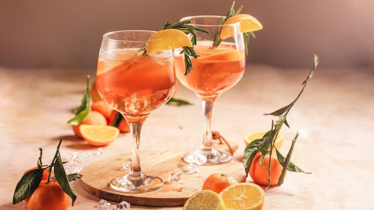 Zwei Aperol Lemon Spritz auf einem hellen Tisch mit Zitronen und Orange. Alles von der Seite fotografiert.