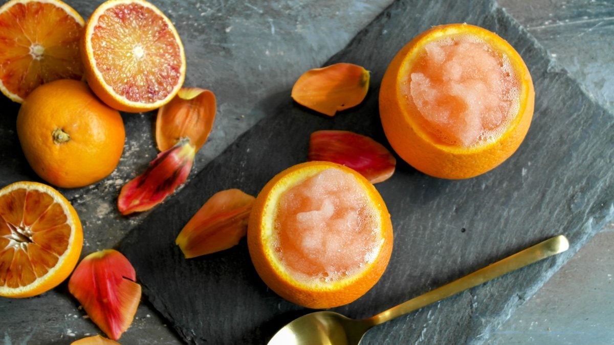 Zwei Orangen mit dem Eis auf einer schwarzen Schiefertafel und grauem Tisch. Ringsherum halbierte Orangen und rötliche Blütenblätter. Alles direkt von oben fotografiert.