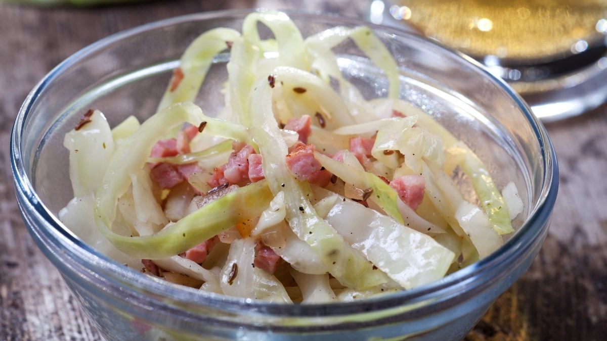 Ein sehr nahes Foto einer kleinen Glasschüssel mit dem Krautsalat auf einem rustikalen Holztisch von leicht schräg oben fotografiert. Im Hintergrund noch unscharf etwas Öl und Weißkohl erkennbar.