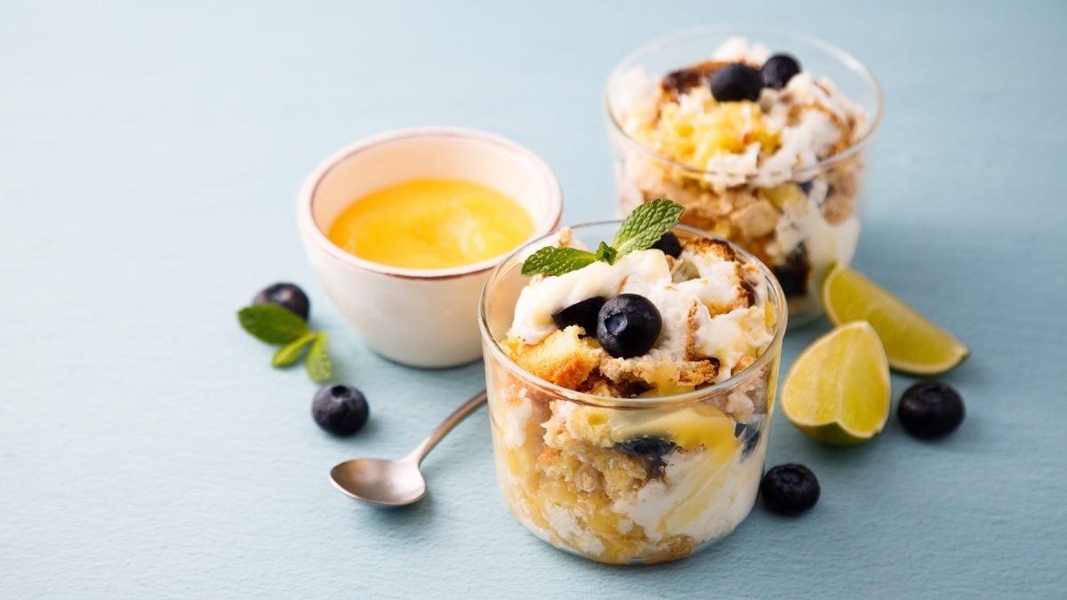 Heidelbeer-Zitronen-Trifle in zwei Gläsern vor blauem Hintergrund, daneben ein Schälchen mit Lemon Curd.