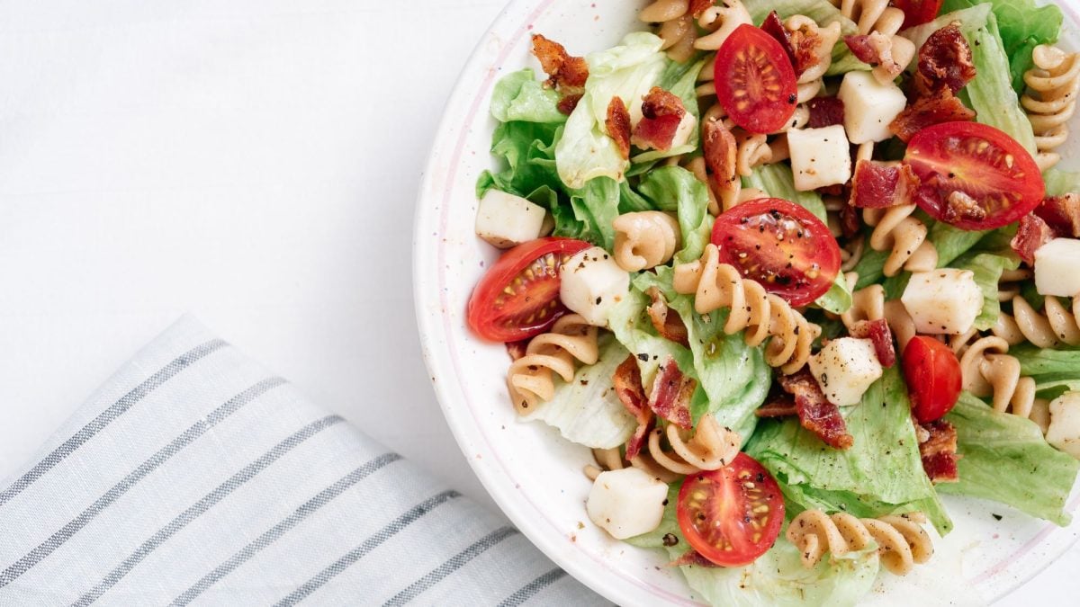 Frischer Nudelsalat mit Tomaten, Salatblättern, Käsewürfeln und Speck auf weißem Hintergrund, daneben ein gestreiftes Tuch.