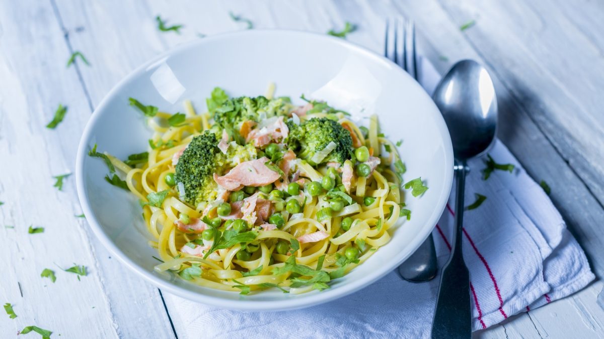 Draufsicht, schräg: Auf hellblauem Grund, eine runde, hellblaue Schüssel Brokkoli-Nudeln mit Erbsen und Lachs.