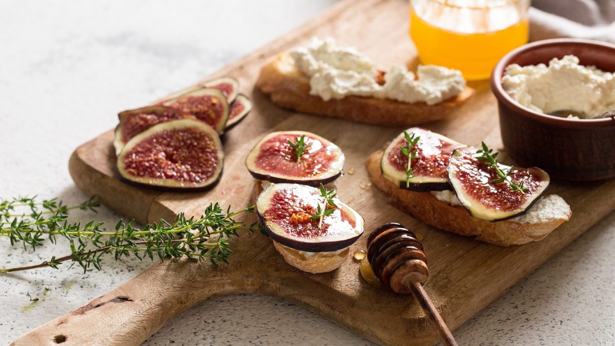 Bruschette mit Ziegenkäse, Honig und Feigen auf einem Holzbrett, davor Honiglöffel.