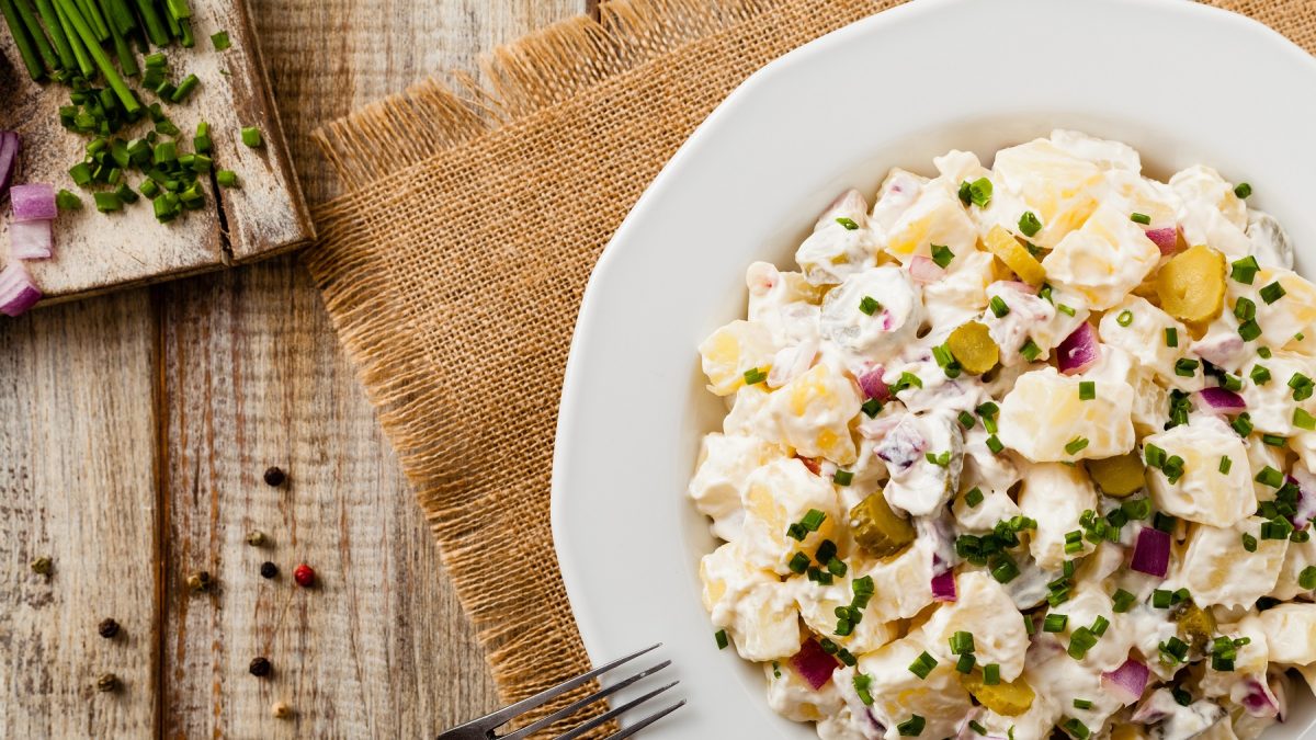 Ein weißer Teller mit dem DDR-Kartoffelsalat direkt von oben auf rustikalem Holztisch, mit Holzbrett fotografiert. Oben links im Bild frischer Schnittlauch, Zwiebel und Pfeffer.