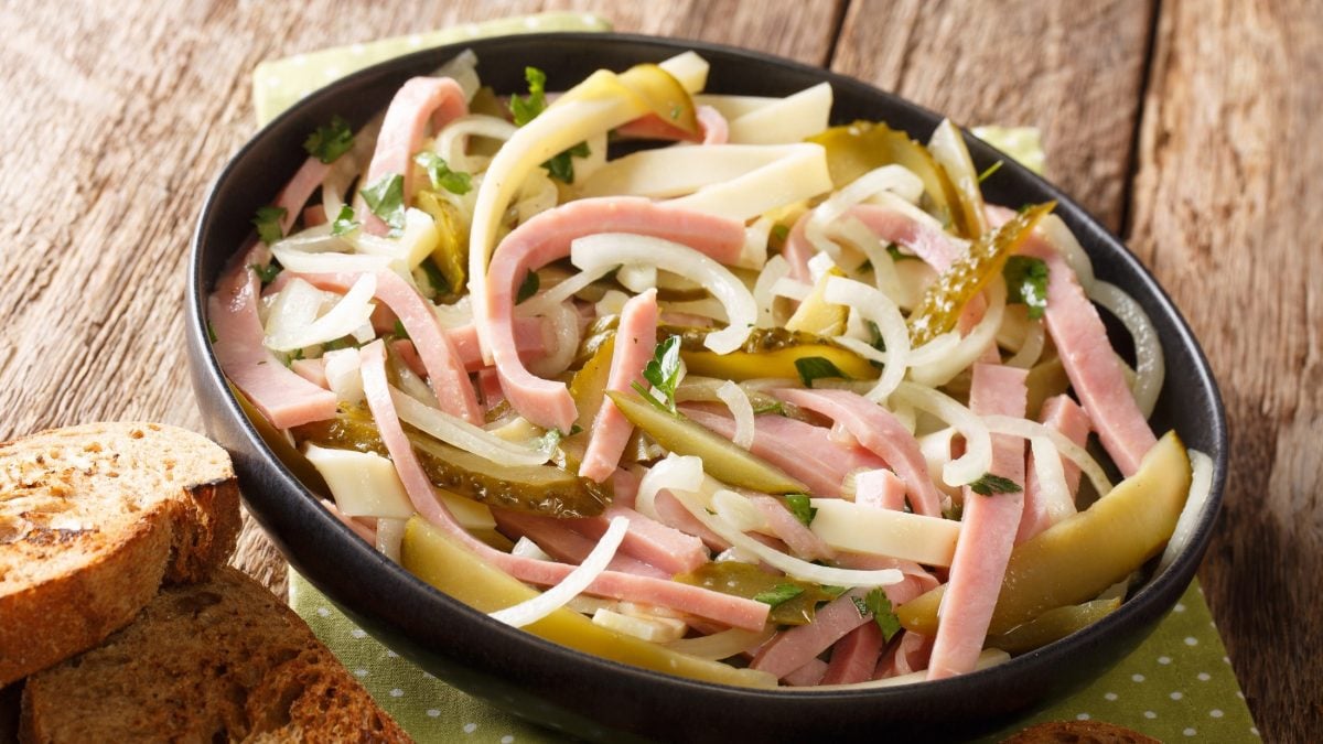 Draufsicht, schräg: Eine runde, dunkle Schale mit einem Elsässer Wurstsalat. Die steht auf einem rustikalen Holzboden.