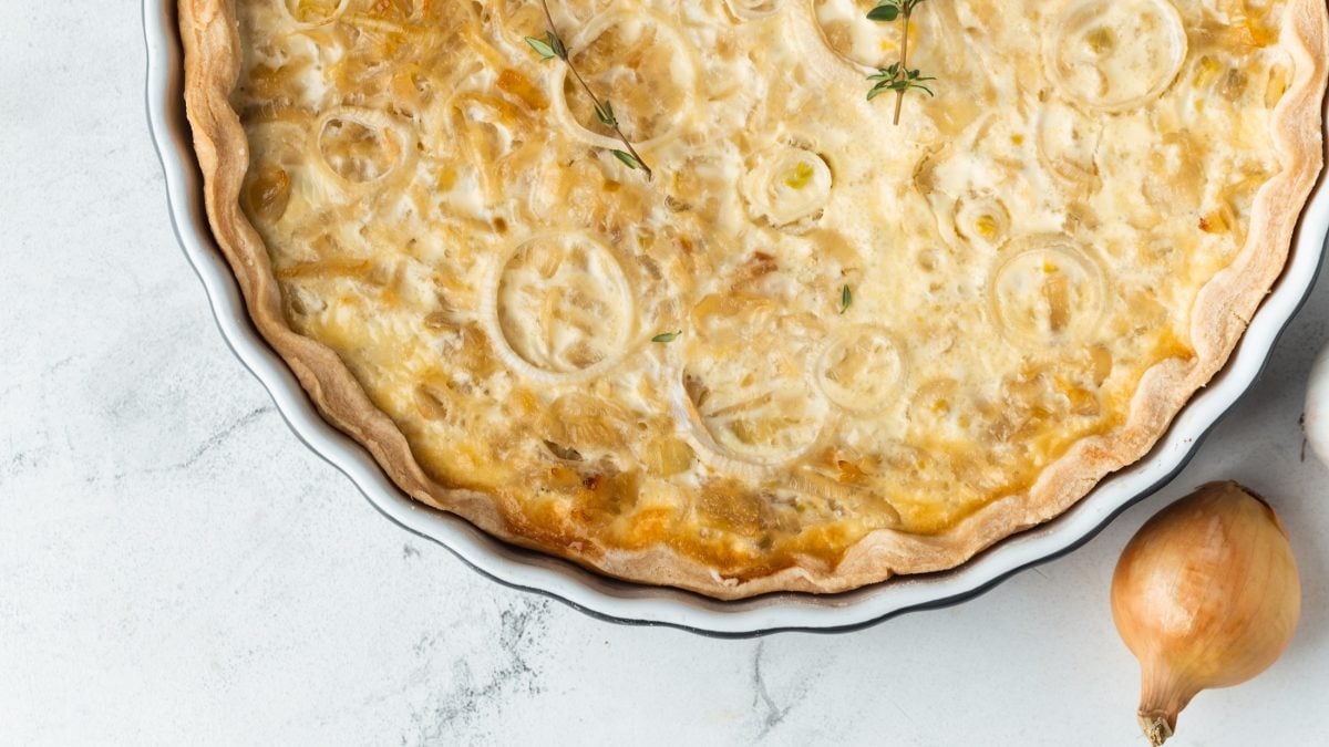 Draufsicht: Auf hellem Marmorboden ist die Hälfte einer runden Form mit einem Elsässer Zwiebelkuchen zu sehen.