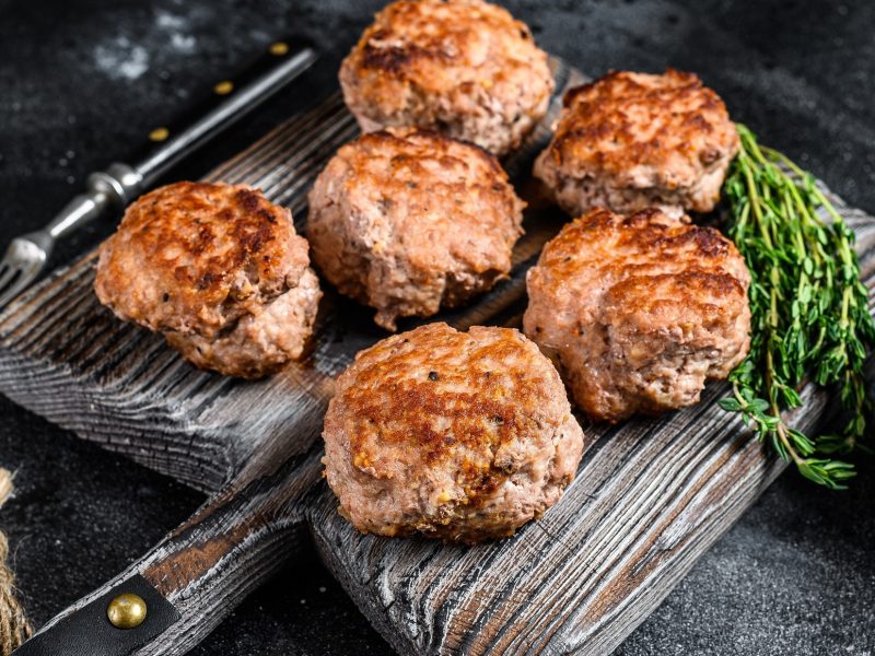 Sechs Frikadellen auf einem rustikalen Holzbrettchen. Die besten Saucen zu Frikadellen. Draufsicht, schräg.