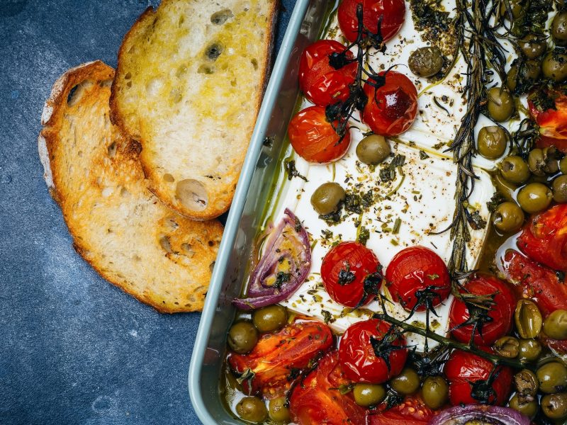 Draufsicht: Auf blauem Untergrund ist eine Auflaufform mit gebackenes Gemüse mit Feta. Es handelt sich um Tomaten, rote Zwiebeln, Oliven und Kräutern.