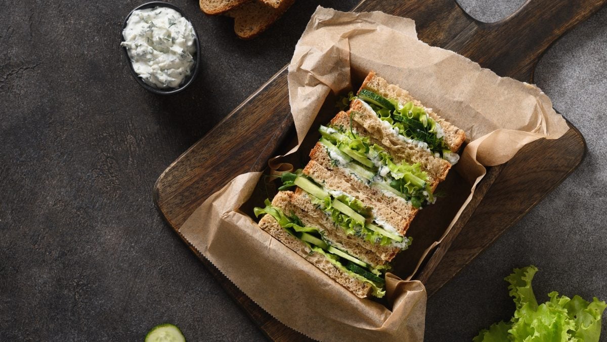 Draufsicht: Fünf verpackte Gurken-Sandwich mit Käse in einer offenen Schachtel, die mit Papier ausgelegt ist. Die Kiste steht auf einem schwarzem Untergrund.