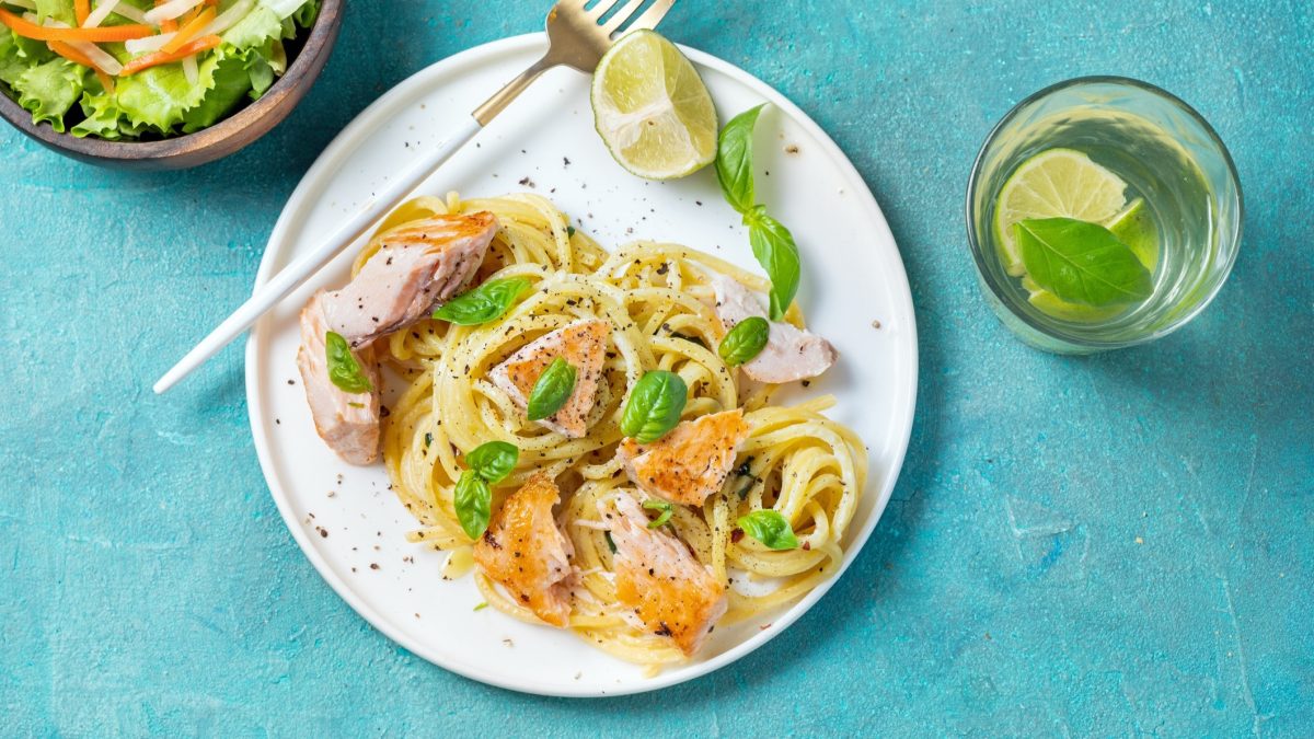 Ein weißer Teller Lachs-Nudeln in Limettensauce auf einem leicht türkises, blauem Untergrund. Daneben sind eine kleine Schüssel mit Salat und ein Glas Wasser mit einer Limette.