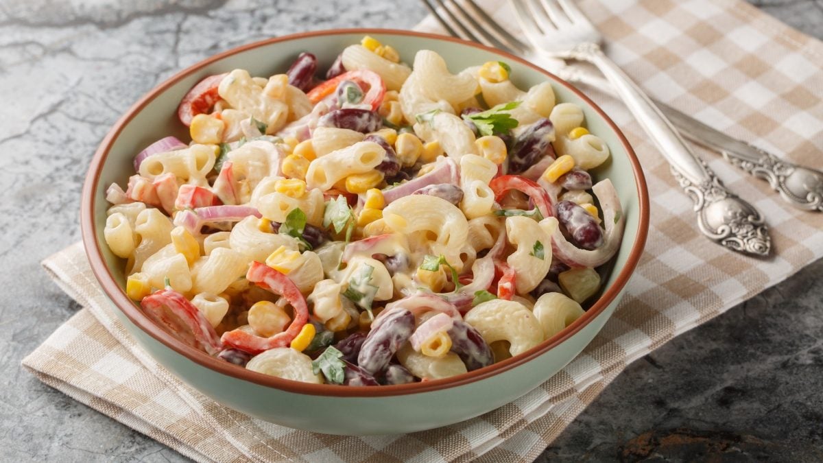 Draufsicht, schräg: Eine rustikale Schale Nudelsalat mit Frischkäse steht auf einem braun-weißes kariertem Tuch. Daneben liegen zwei gabeln auf Silber.