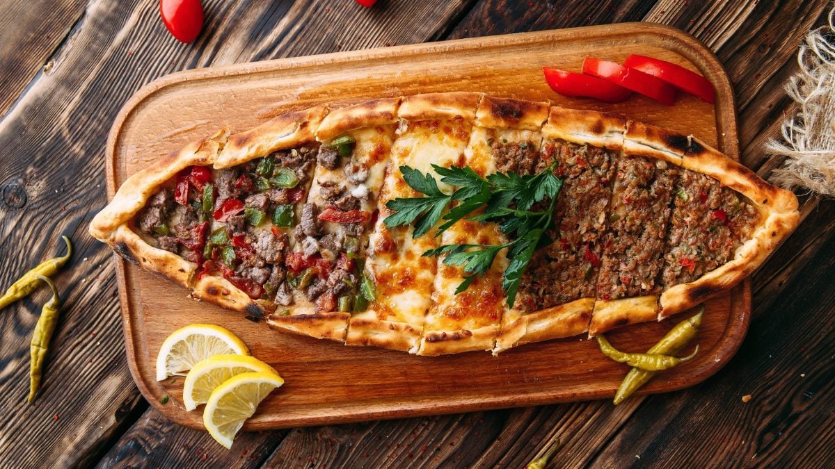 Draufsicht: Ein Holzbrett mit einer selbst gemachten türkischen Pide mit Hackfleisch. Das liegt auf einem Holzboden.