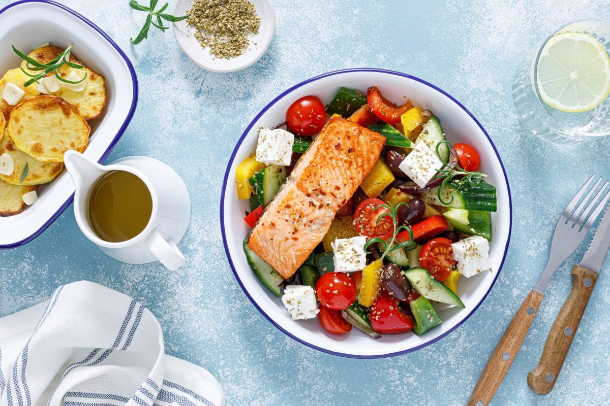 Draufsicht: Ein mediterraner Sommersalat mit Lachsfilet auf einem hellblauen Untergrund. Daneben liegt ein Besteck. Eine weitere Schüssel mit Bratkartoffeln und ein Kännchen mit Olivenöl sind ebenfalls zu sehen.
