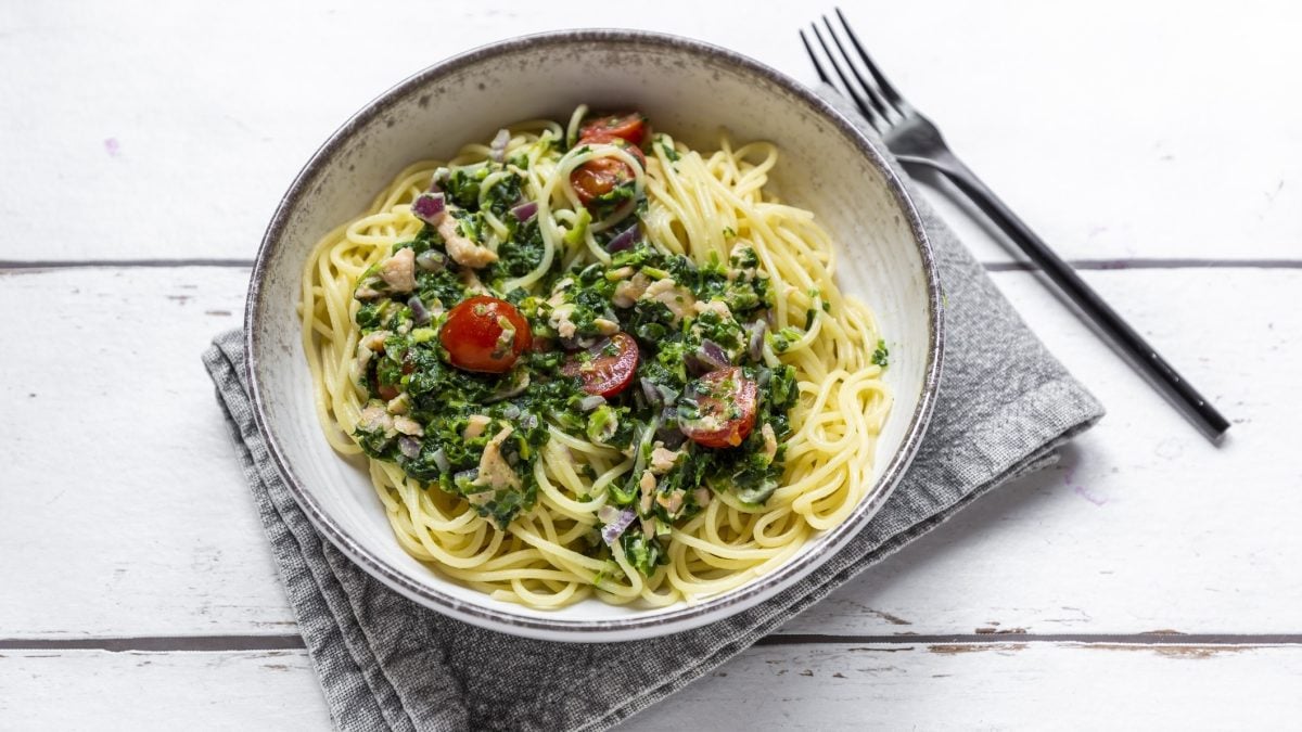 Draufsicht: Eine runde weiße Schüssel mit Spaghetti in Spinat-Lachs-Sauce auf einem weißen Untergrund aus Holz. Die Schüssel steht auf einem grauen Tuch. Daneben liegt eine Gabel.