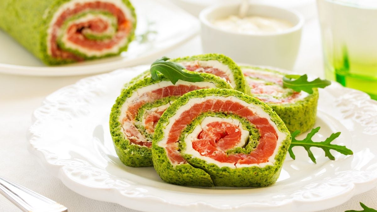 Frontal: Ein weißer Teller mit drei Scheiben einer aufgeschnittenen Spinat-Lachs-Roulade. Im Hintergrund ist die restliche Spinat-Lachs-Rolle.
