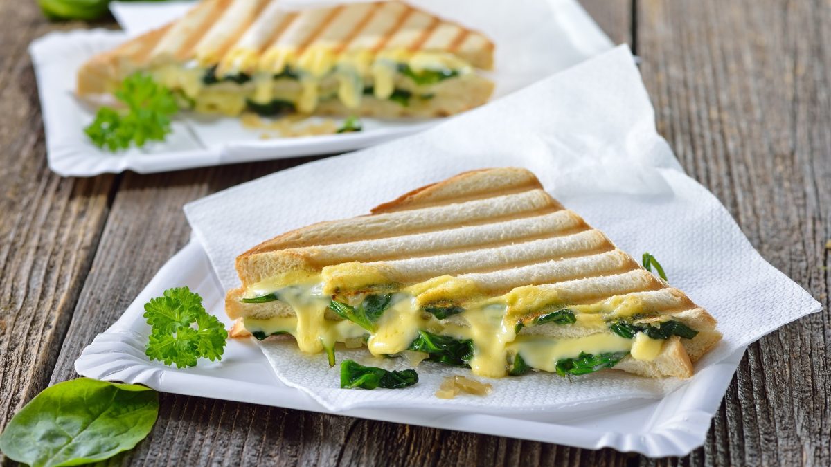 Zwei diagonal geschnittene Spinat-Tramezzini mit Käse auf einer weißen Papierserviette. Die liegen auf einem Untergrund auf grobem Holz.