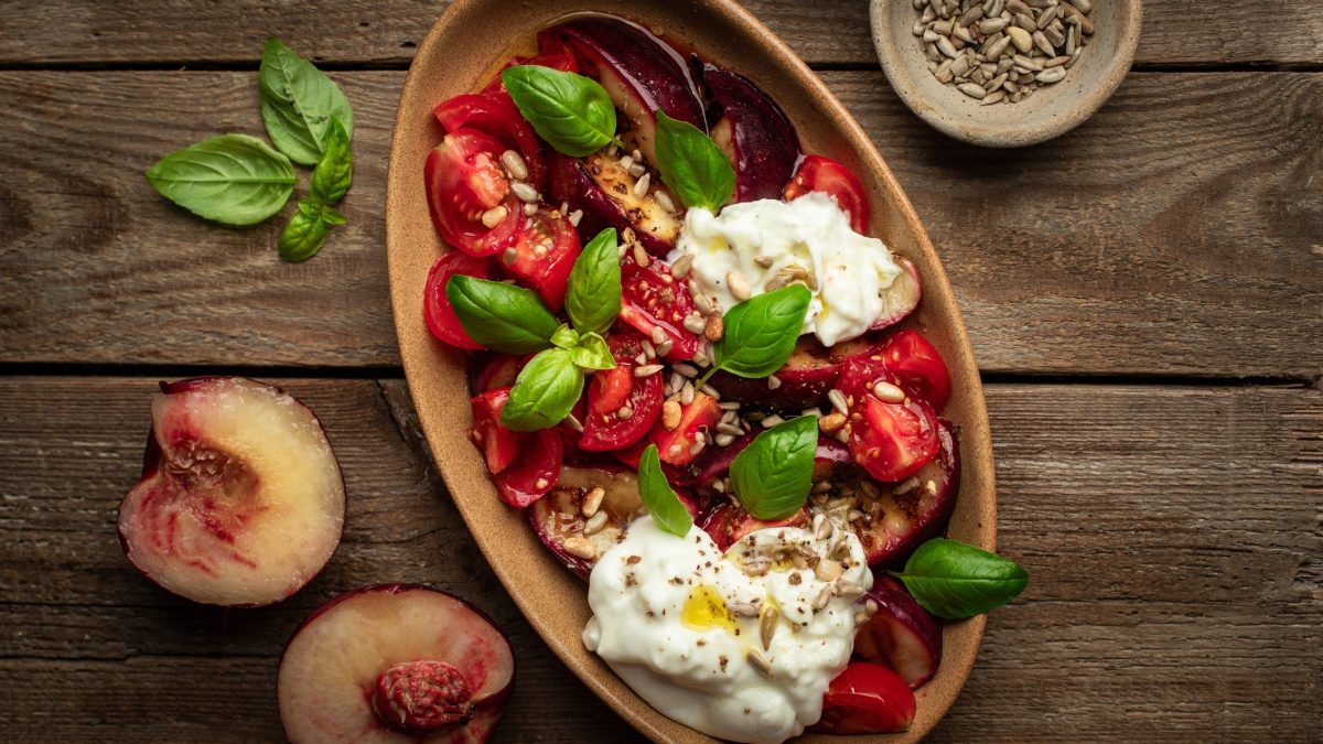Draufsicht: Ein Tomaten-Pfirsich-Salat mit Burrata in einer ovalen Form. Die steht auf einem Untergrund aus rustikalen Holzplanken. Daneben liegen Pfirsich-Hälften und Basilikumblätter.
