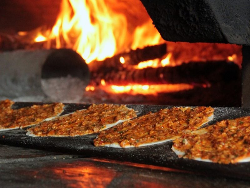 Frontal: Vier Fladen mit Hackfleisch werden in einen offenen Ofen, in dem ein Feuer brennt, geschoben. Der Fladen wurde nach Türkische Hackfleisch-Rezept zubereitet,