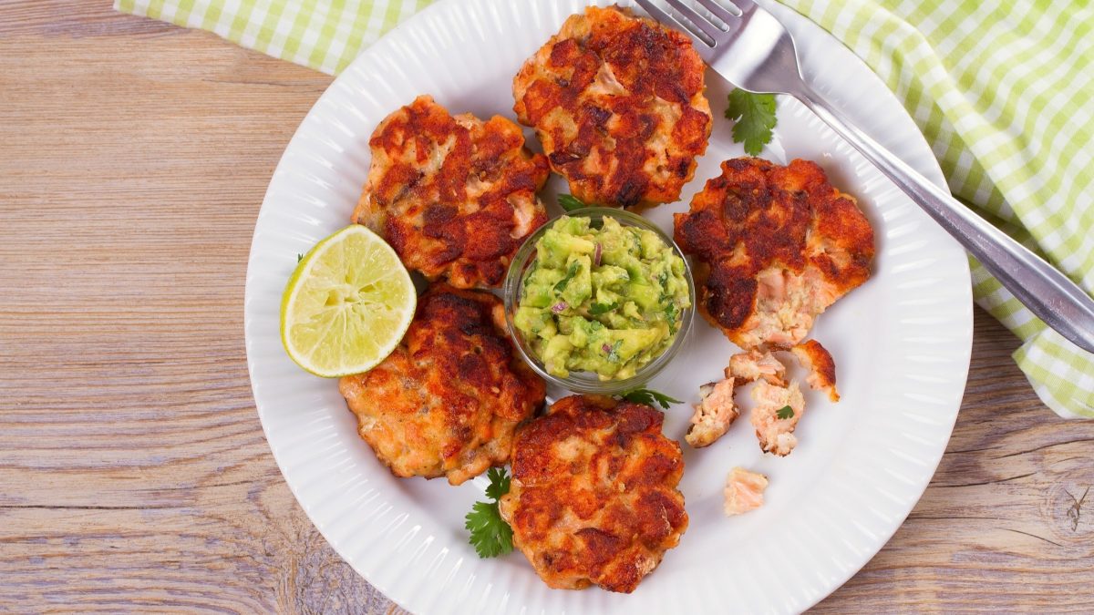 Draufsicht: Selbst gemachte Lachs-Patties auf einem weißen Teller. In der Mitte steht ein Glasschälchen mit Avocado-Creme. Auf dem teller liegt eine Gabel. Und neben dem Teller ein hellgrün und weiß kariertes Handtuch.