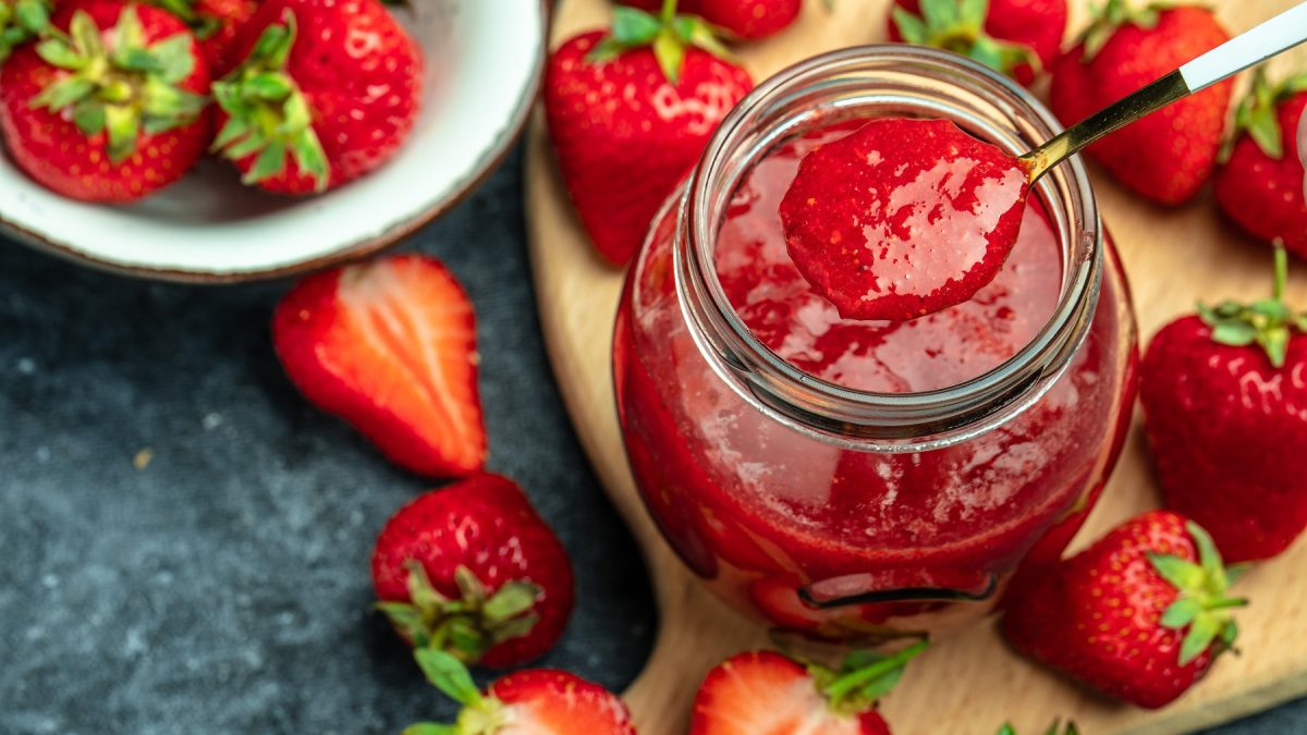 Ein Löffel hebt Erdbeermarmelade aus einem Glas, umgeben von reifen Erdbeeren auf einem dunklen Untergrund.