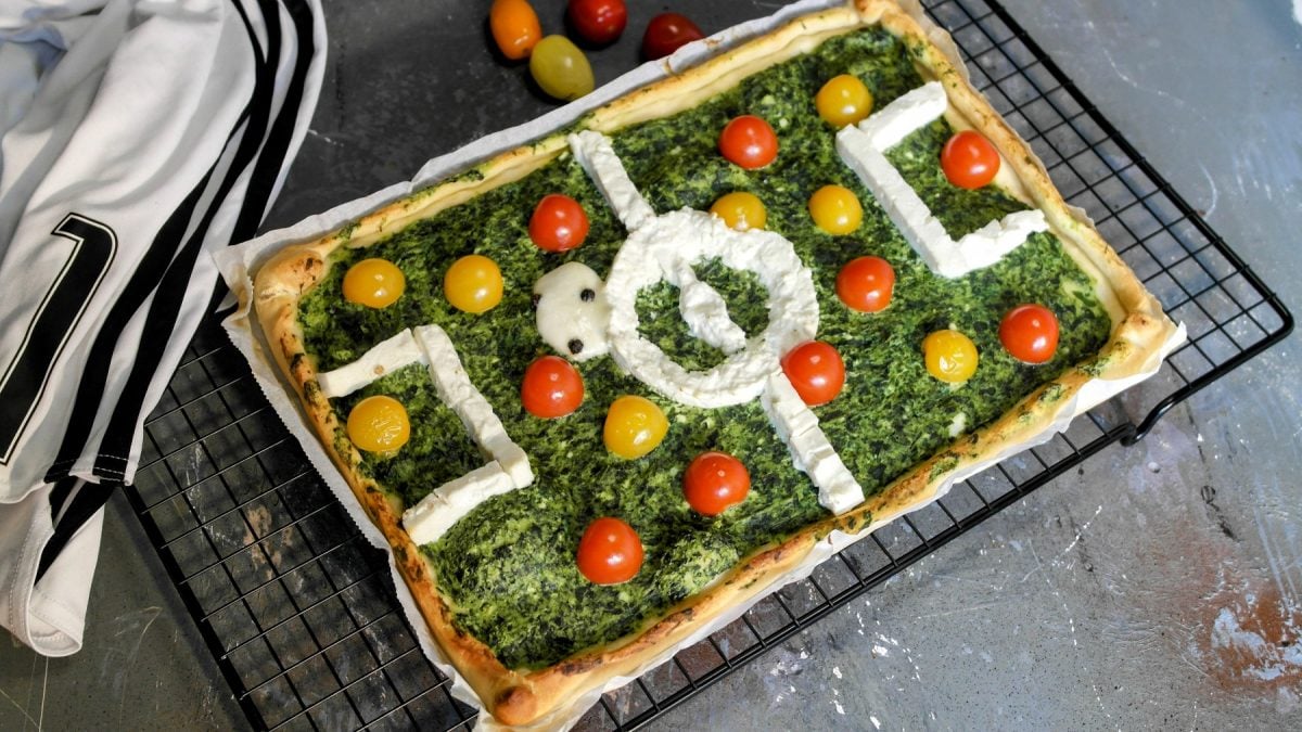Die Fussball-Pizza auf einem schwarzen Metallgitter und grauem Steintisch. Daneben ein paar bunte Kirschtomaten und eine weiß-schwarze Fußball-Shorts. Alles von leicht schräg oben fotografiert.