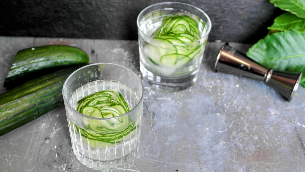 Zwei Gläser mit den Gurken-Eiswürfeln auf einem Steintisch mit schwarzem Hintergrund.