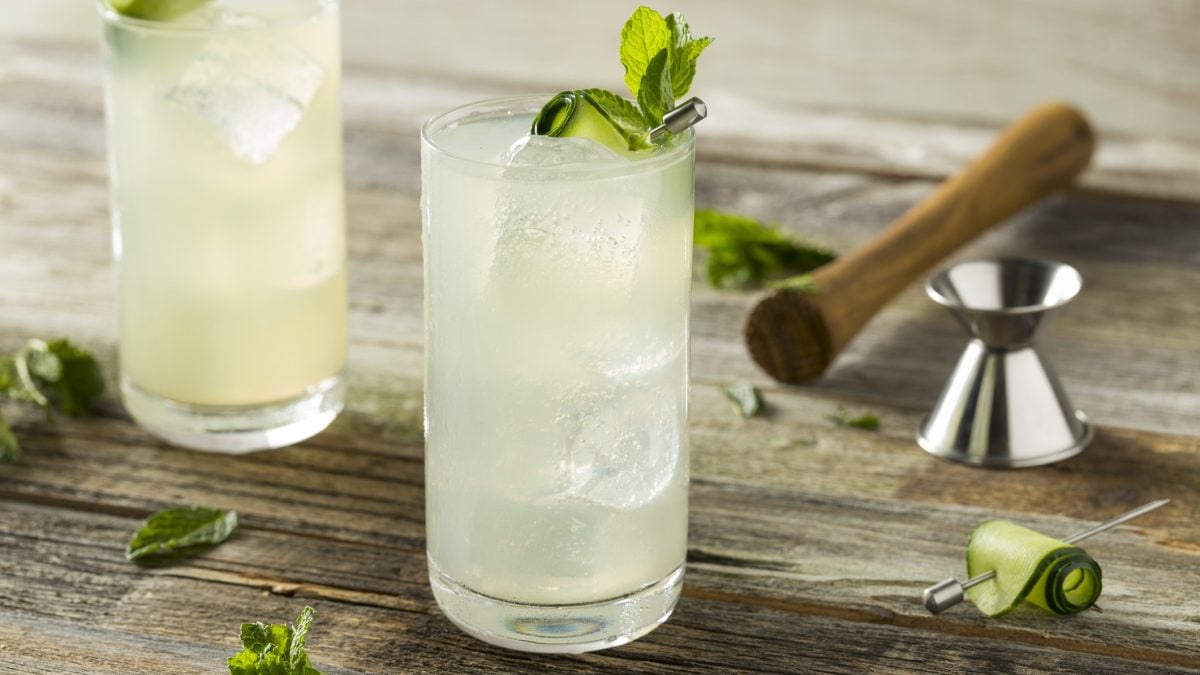 Zwei Gläser mit dem Gurken-Spritz auf einem rustikalen Holztisch mit Minze, Gurke und Barzubehör. Alles von der Seite fotografiert.