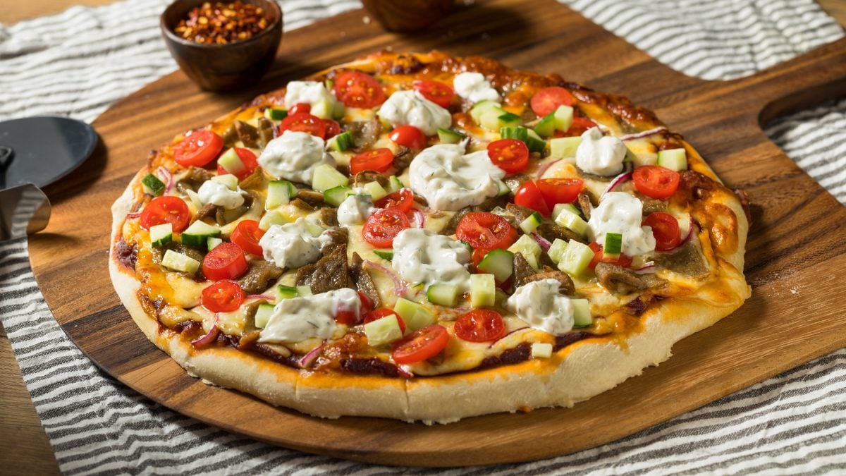 Ein großes Holzbrett mit der Pizza auf einem Tisch mit grau-weiß-gestreifter Tischdecke. Alles von schräg oben fotografiert.