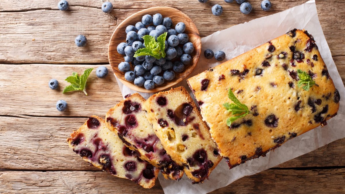 Frisch gebackener Heidelbeer-Mascarpone-Kuchen mit Minzblättern auf Backpapier, daneben eine Schale Blaubeeren auf rustikalem Holztisch.