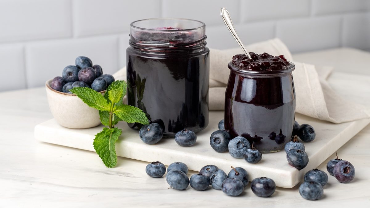 Gläser mit dunkler Heidelbeermarmelade und frische Blaubeeren auf weißem Brett in heller Küche arrangiert.