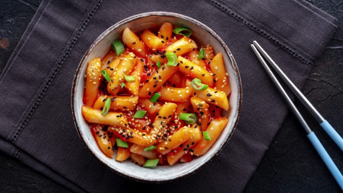 Honig-Butter-Tteokbokki in einer Schüssel auf schwarzem Hintergrund. Rechts auf dem Bild Stäbchen.
