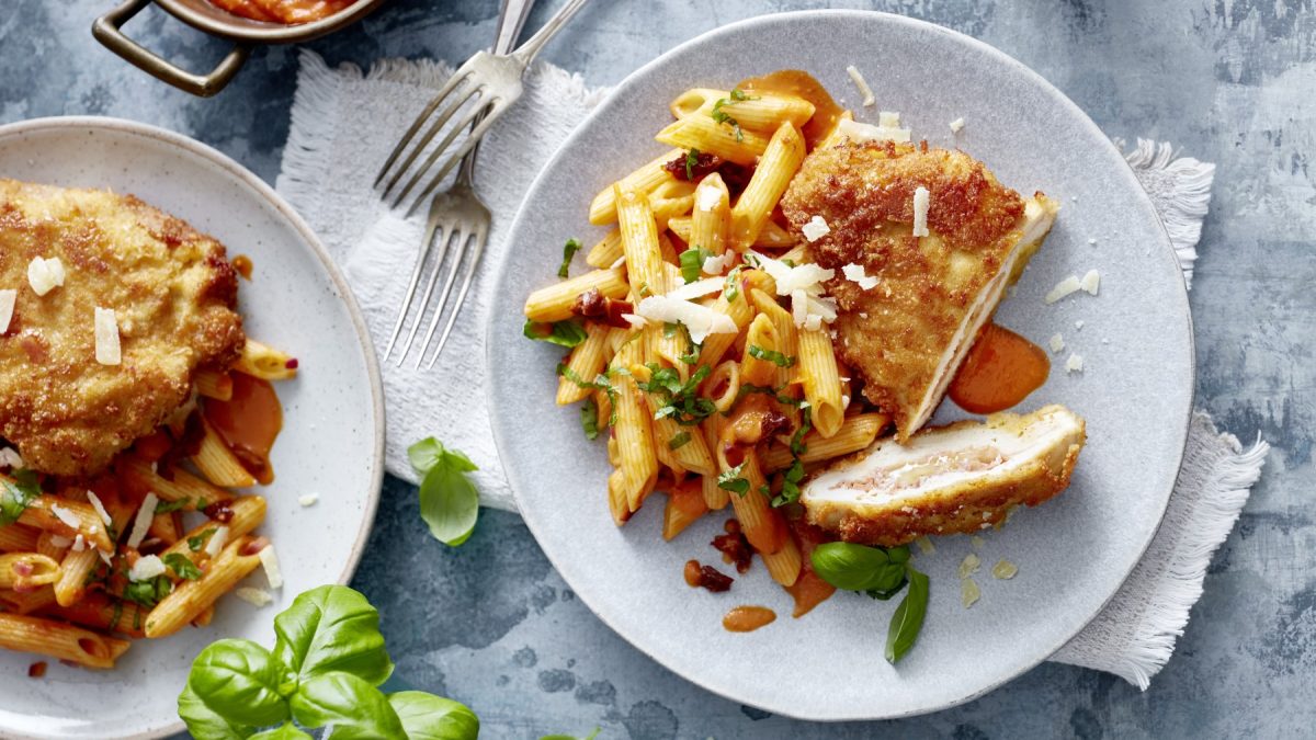 Draufsicht: Italienisches Hähnchen-Cordon-bleu auf je zwei Tellern, daneben Basilikum und Gabeln, grauer Untergrund.