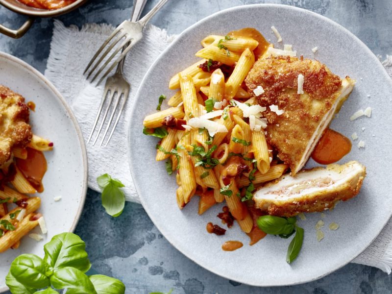 Draufsicht: Italienisches Hähnchen-Cordon-bleu auf je zwei Tellern, daneben Basilikum und Gabeln, grauer Untergrund.
