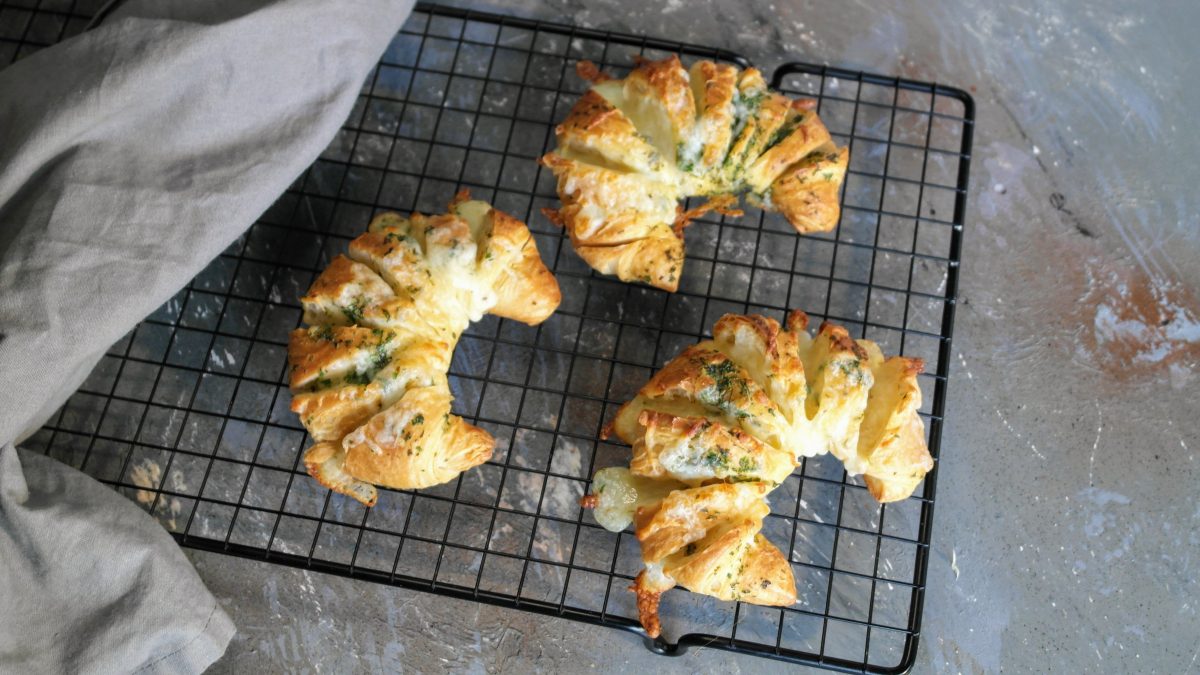 Drei Käse-Kräuter-Croissants auf einem schwarzen Gitter und grauem Untergrund direkt von oben fotografiert.
