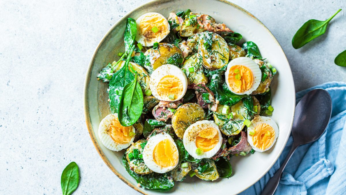Eine weiße Schale mit dem Kartoffelsalat auf grau-blauem Untergrund und einer blauen Tischdecke mit silbernem Löffel unten links. Alles direkt von oben fotografiert.