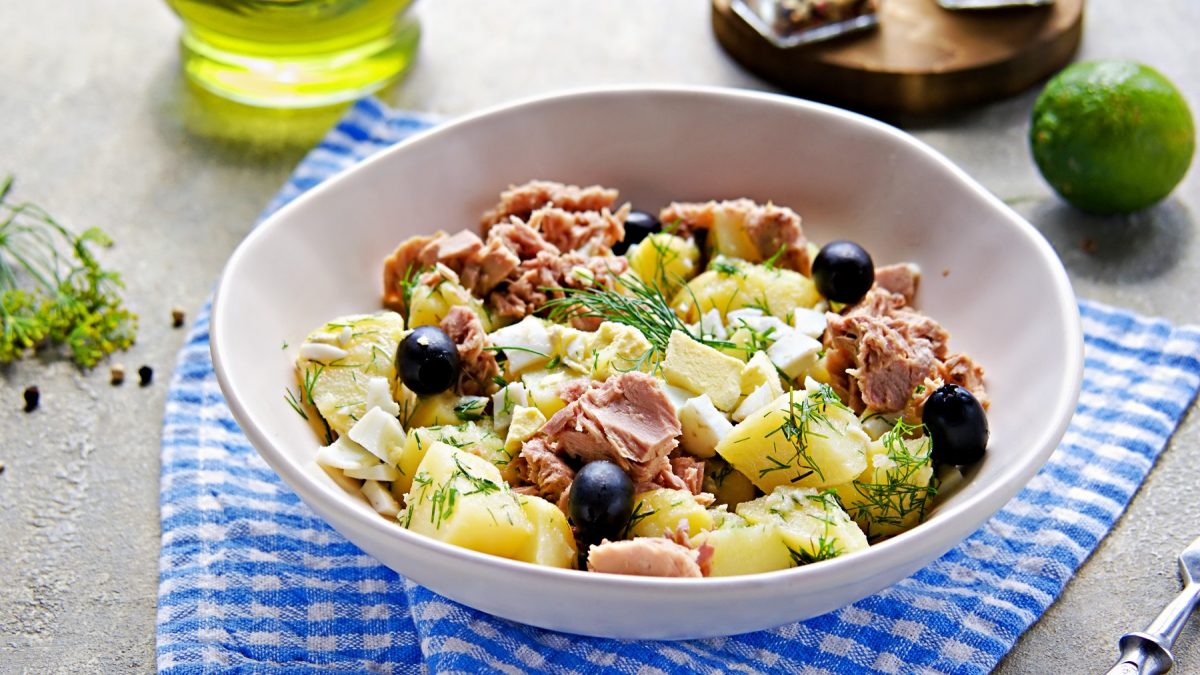 Eine weiße Schale mit dem Kartoffelsalat auf einem blau-weiß-gestreiftem Tuch und dunklem Holztisch. Alles von schräg oben fotografiert.