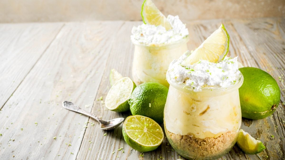 Zwei Gläser mit dem Key Lime Pie auf einem rustikalen Holztisch mit frischen Limetten und einem kleinen Löffel aus Metall. Alles von der Seite fotografiert.